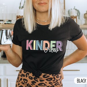 May include: A black t-shirt with the text "Kinder Crew" in rainbow colors. The text is in a playful font. The shirt is being worn by a person with long blonde hair. The person is holding a black and white mug.