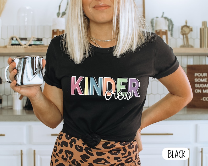 May include: A black t-shirt with the text "Kinder Crew" in rainbow colors. The text is in a playful font. The shirt is being worn by a person with long blonde hair. The person is holding a black and white mug.