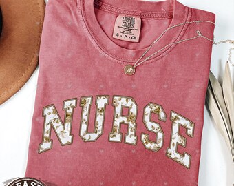May include: A crimson-colored t-shirt with the word "NURSE" in a floral design. The shirt has a crew neck and short sleeves. A silver necklace with a pendant is draped across the shirt. The shirt is labeled "Comfort Colors".