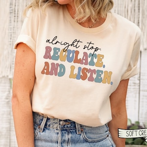 May include: A beige t-shirt with the text "alright stop. REGULATE. AND LISTEN." in colorful, retro-style lettering.