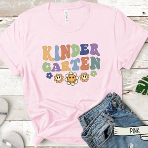 Puede incluir: Camiseta rosa con el texto "KINDERGARTEN" en colores arco&iacute;ris y un dise&ntilde;o floral. La camiseta est&aacute; hecha de un material suave y c&oacute;modo.