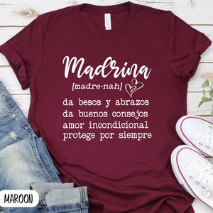 May include: A maroon t-shirt with white text that reads "Madrina [madre-nah] da besos y abrazos da buenos consejos amor incondicional protege por siempre"  MAROON
