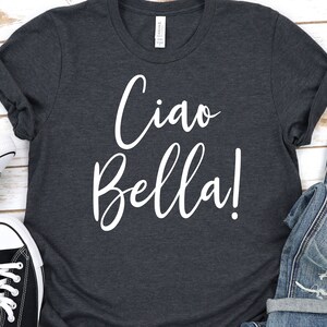 K&ouml;nnte beinhalten: Dunkelgraues T-Shirt mit den Worten "Ciao Bella!" in wei&szlig;er Schrift. Das Shirt hat hochgekrempelte &Auml;rmel und wird mit schwarz-wei&szlig;en Turnschuhen und Jeans kombiniert.