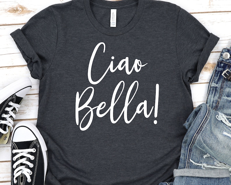 K&ouml;nnte beinhalten: Dunkelgraues T-Shirt mit den Worten "Ciao Bella!" in wei&szlig;er Schrift. Das Shirt hat hochgekrempelte &Auml;rmel und wird mit schwarz-wei&szlig;en Turnschuhen und Jeans kombiniert.