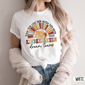 May include: White t-shirt with a rainbow and flower design. The text "Teach them, love them, watch them grow, Kindergarten dream team" is printed on the shirt.