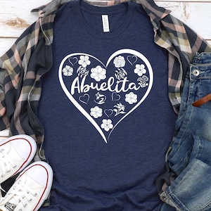 May include: A navy blue t-shirt with a white heart design featuring the word "Abuelita" surrounded by flowers. The shirt is laid out on a wooden surface with a plaid shirt and blue jeans.