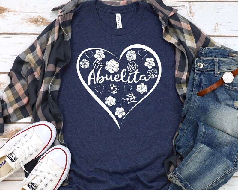 May include: A navy blue t-shirt with a white heart design featuring the word "Abuelita" surrounded by flowers. The shirt is laid out on a wooden surface with a plaid shirt and blue jeans.