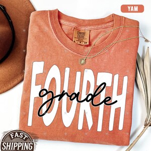 Op de afbeelding: Een koraal gekleurd T-shirt met de woorden "FOURTH grade" in witte en zwarte letters.