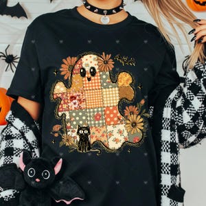 May include: Black t-shirt featuring a patchwork ghost design with floral and polka dot patterns. A black cat sits below the ghost. The design includes flowers and a bat, perfect for Halloween.