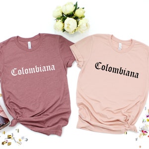 Op de afbeelding: Twee T-shirts met het woord "Colombiana" gedrukt in een gotisch lettertype. Een shirt is stoffig roze en de andere is lichtroze.