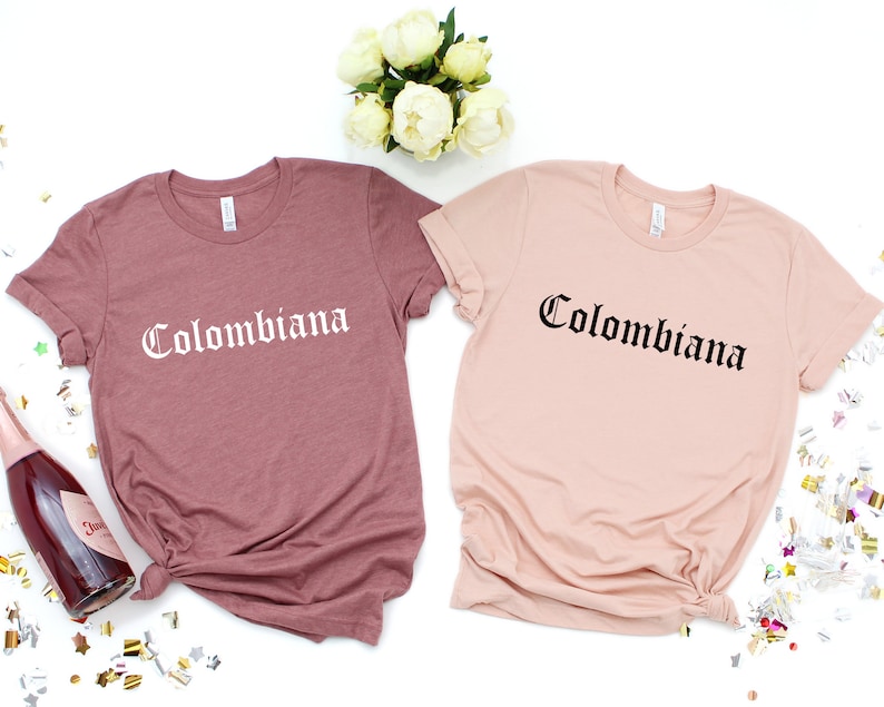 Op de afbeelding: Twee T-shirts met het woord "Colombiana" gedrukt in een gotisch lettertype. Een shirt is stoffig roze en de andere is lichtroze.