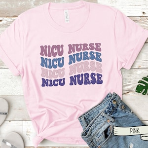 May include: Pink t-shirt with the text "NICU NURSE" repeated four times in a retro font. The text is in shades of purple, blue, and pink.