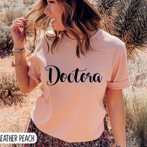 K&ouml;nnte beinhalten: Ein rosa T-Shirt mit dem Wort "Doctora" in schwarzer, kursiv geschriebener Schrift. Das Shirt wird von einer Person in einer W&uuml;stenlandschaft getragen.