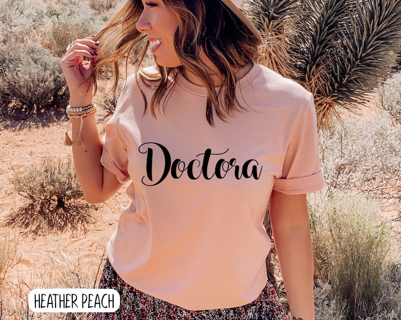 K&ouml;nnte beinhalten: Ein rosa T-Shirt mit dem Wort "Doctora" in schwarzer, kursiv geschriebener Schrift. Das Shirt wird von einer Person in einer W&uuml;stenlandschaft getragen.