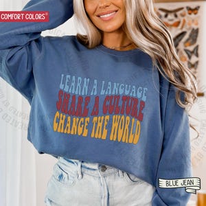 May include: A blue sweatshirt with the text "LEARN A LANGUAGE, SHARE A CULTURE, CHANGE THE WORLD" in retro font. The sweatshirt is a Comfort Colors brand and is paired with blue jeans.