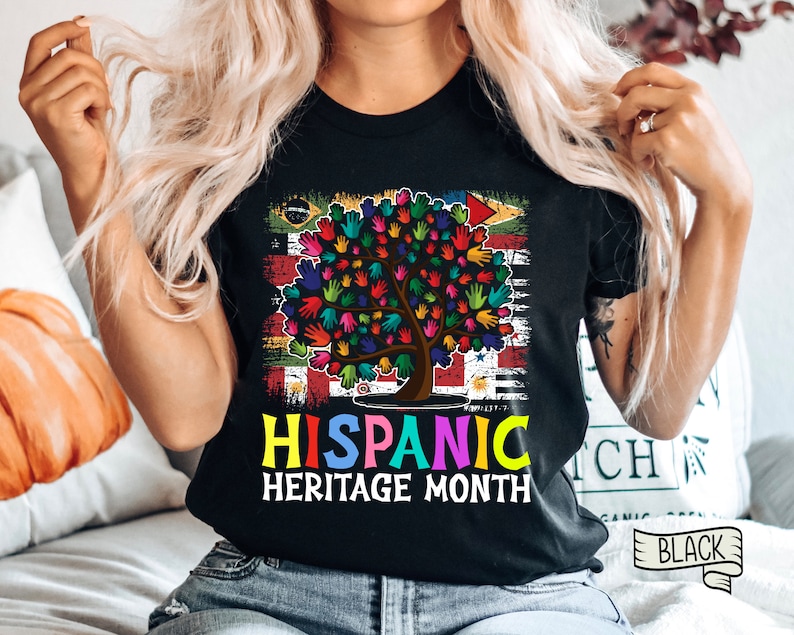 May include: Black t-shirt with a colorful tree made of handprints and the text "Hispanic Heritage Month". The tree is surrounded by a flag with different colors and designs.