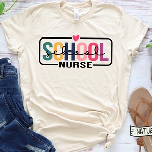 May include: A beige t-shirt with the text "SCHOOL NURSE" in a colorful block font with a pink heart above the text. The text is framed in a black rectangle.