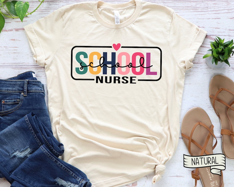 May include: A beige t-shirt with the text "SCHOOL NURSE" in a colorful block font with a pink heart above the text. The text is framed in a black rectangle.