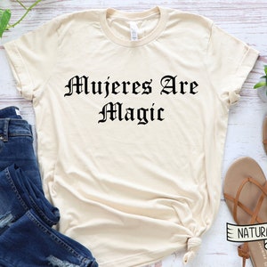 May include: A beige t-shirt with the text "Mujeres Are Magic" printed in black in a gothic font.