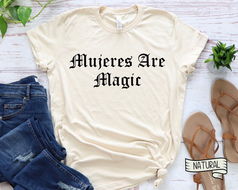 May include: A beige t-shirt with the text "Mujeres Are Magic" printed in black in a gothic font.