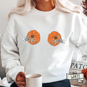Op de afbeelding: Witte trui met een afbeelding van twee oranje pompoenen die door skelethandjes worden vastgehouden. De tekst "White" staat op een wit lint.