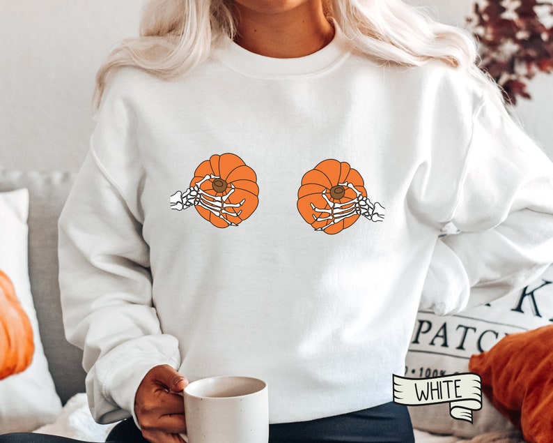 Op de afbeelding: Witte trui met een afbeelding van twee oranje pompoenen die door skelethandjes worden vastgehouden. De tekst "White" staat op een wit lint.
