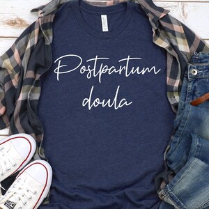 May include: A navy blue t-shirt with the text "Postpartum Doula" in white script lettering. The shirt is laid flat on a white wooden surface with a plaid shirt and blue jeans.