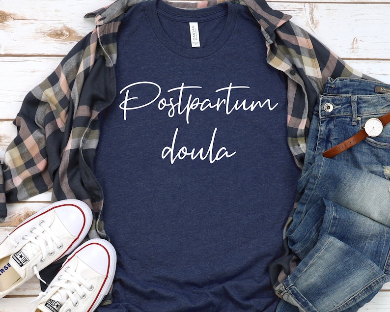 May include: A navy blue t-shirt with the text "Postpartum Doula" in white script lettering. The shirt is laid flat on a white wooden surface with a plaid shirt and blue jeans.