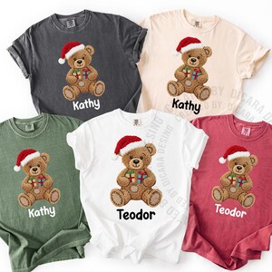 May include: A collection of five t-shirts in various colors, each featuring a Christmas teddy bear design. The bear wears a Santa hat and holds wrapped gifts. The names "Kathy" and "Teodor" are printed below the bear.