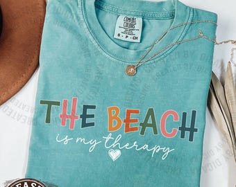 Può includere: Una t-shirt a maniche corte color verde schiuma di mare con scollo rotondo. Il capo presenta la scritta THE BEACH in lettere stampatello multicolori e il testo in corsivo is my therapy con una piccola icona a forma di cuore bianco sotto.