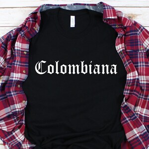 Op de afbeelding: Zwart T-shirt met het woord "Colombiana" in wit, in gotische letters. Het shirt ligt op een rood-wit geruit flanellen overhemd.