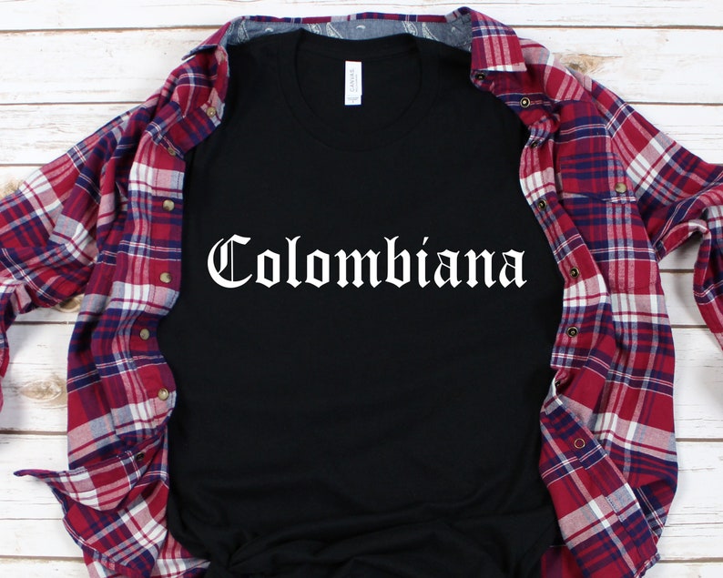 Op de afbeelding: Zwart T-shirt met het woord "Colombiana" in wit, in gotische letters. Het shirt ligt op een rood-wit geruit flanellen overhemd.
