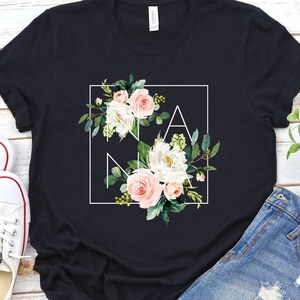 May include: Black t-shirt featuring a white square frame enclosing the word "NANA" in white lettering. The letters are surrounded by pink and white flowers and green leaves.