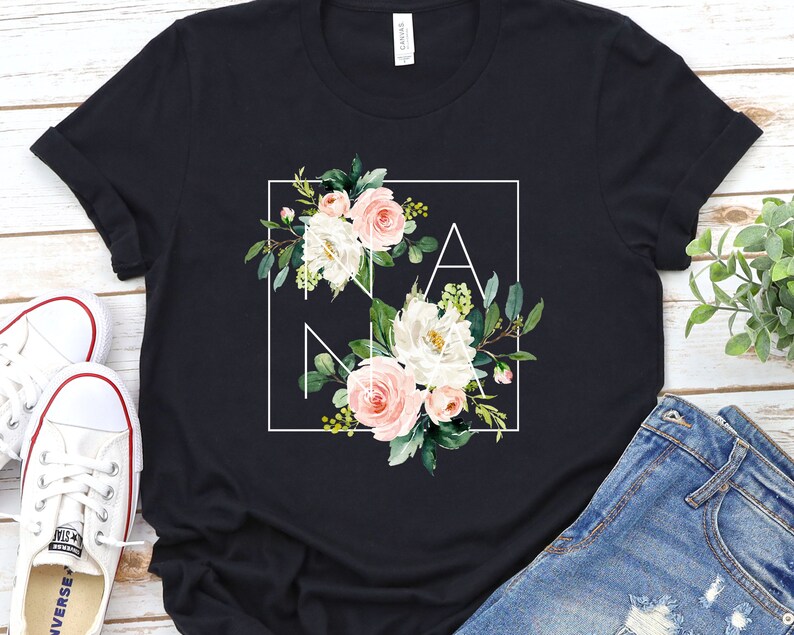 May include: Black t-shirt featuring a white square frame enclosing the word "NANA" in white lettering. The letters are surrounded by pink and white flowers and green leaves.