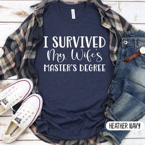 May include: A heather navy blue t-shirt with the text "I survived my wife's master's degree" in white. The shirt is displayed on a wooden surface with a pair of white sneakers, a plaid shirt, and blue jeans.