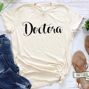 K&ouml;nnte beinhalten: Ein beiges T-Shirt mit dem Wort "Doctora" in schwarzer Schrift.