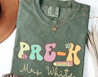 Peut inclure: Un t-shirt vert avec le texte "PRE-K Mrs. White" en lettres colorées de style rétro. Un crayon et une fleur sont également inclus dans le design.