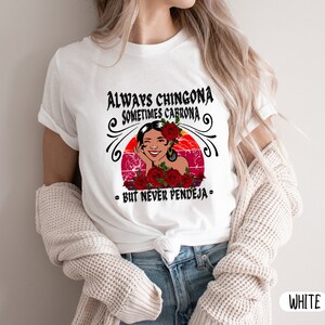 May include: White t-shirt with a graphic design featuring a woman with red flowers and the text "Always Chingona Sometimes Cabrona - But Never Pendeja -"