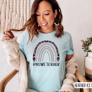 May include: A light blue t-shirt featuring a rainbow design with the text "Daycare Teacher". The rainbow is decorated with pink, green, and brown accents and has a leopard print border.