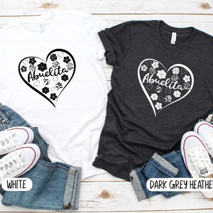 May include: Two t-shirts with a heart design featuring the text "Abuelita" and floral accents. The t-shirts are shown on a white wood background with blue jeans and white sneakers. One t-shirt is white and the other is dark grey heather.
