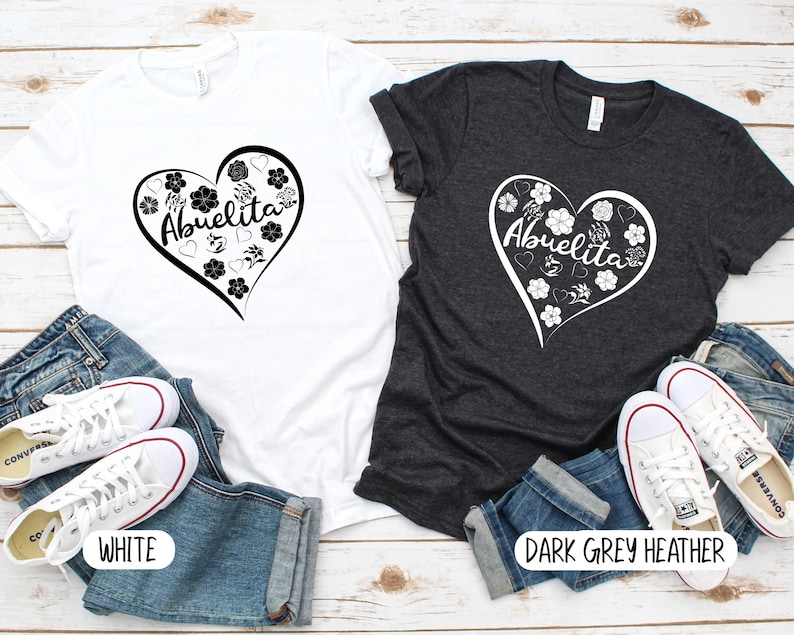 May include: Two t-shirts with a heart design featuring the text "Abuelita" and floral accents. The t-shirts are shown on a white wood background with blue jeans and white sneakers. One t-shirt is white and the other is dark grey heather.