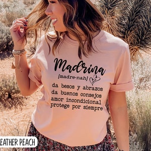 May include: A pink t-shirt with the word "Madrina" in black script, with the definition in Spanish below: "[madre-nah] da besos y abrazos da buenos consejos amor incondicional protege por siempre".