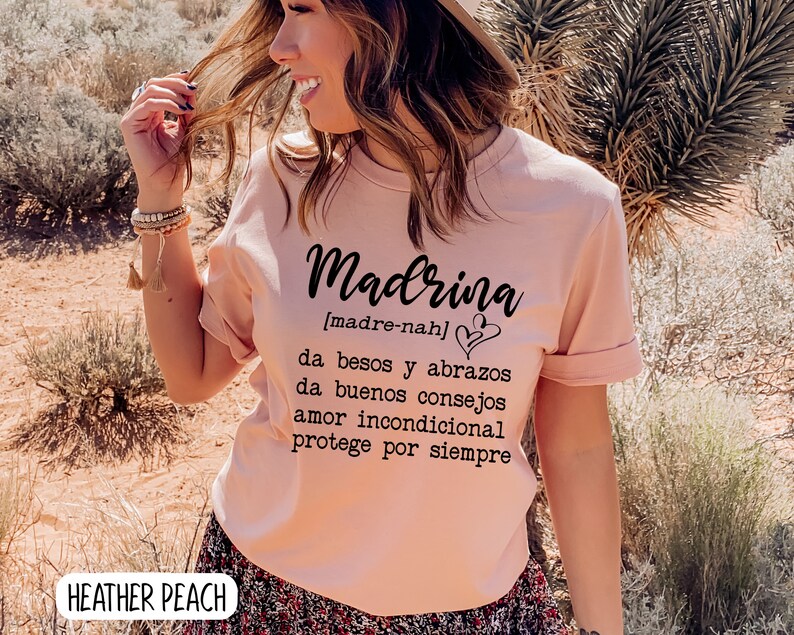 May include: A pink t-shirt with the word "Madrina" in black script, with the definition in Spanish below: "[madre-nah] da besos y abrazos da buenos consejos amor incondicional protege por siempre".