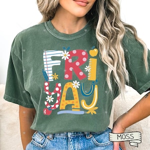 May include: Moss green t-shirt with the word "FRIDAY" in colorful, playful lettering. The design incorporates elements like flowers, an apple, and a ruler, creating a fun, school-themed aesthetic. The shirt is made of a soft material.