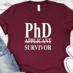 May include: Burgundy t-shirt with white text that reads "PhD Applicant Survivor".