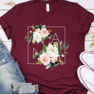 May include: Burgundy t-shirt with a white square frame around a floral design that spells out "NANA".