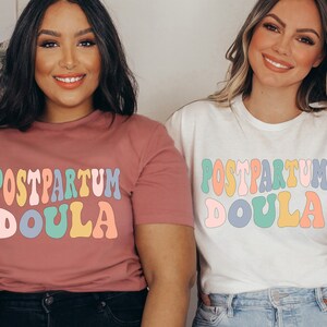 May include: Two women wearing t-shirts with the text "POSTPARTUM DOULA" in colorful retro font. The shirts are pink and white.