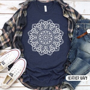 May include: A heather navy blue t-shirt with a white mandala design. The shirt is shown on a model with a plaid shirt, jeans, and white sneakers. The text "HEATHER NAVY" is visible on the bottom right corner of the image.