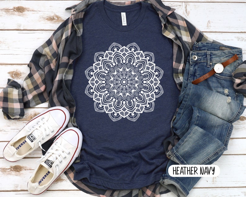 May include: A heather navy blue t-shirt with a white mandala design. The shirt is shown on a model with a plaid shirt, jeans, and white sneakers. The text "HEATHER NAVY" is visible on the bottom right corner of the image.