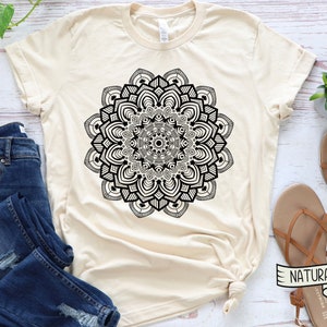 May include: A beige t-shirt with a black mandala design. The shirt is laid flat on a white wooden surface with a pair of brown sandals and a pair of blue jeans.